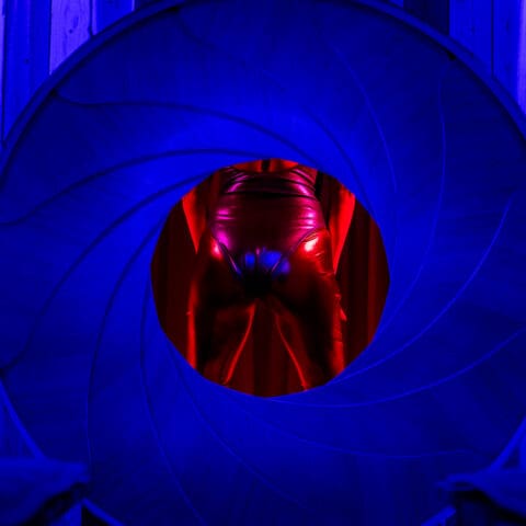 A woman’s backside in a pink shiny leotard cast in red light inside of a round blue shutter.