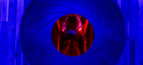 A woman’s backside in a pink shiny leotard cast in red light inside of a round blue shutter.