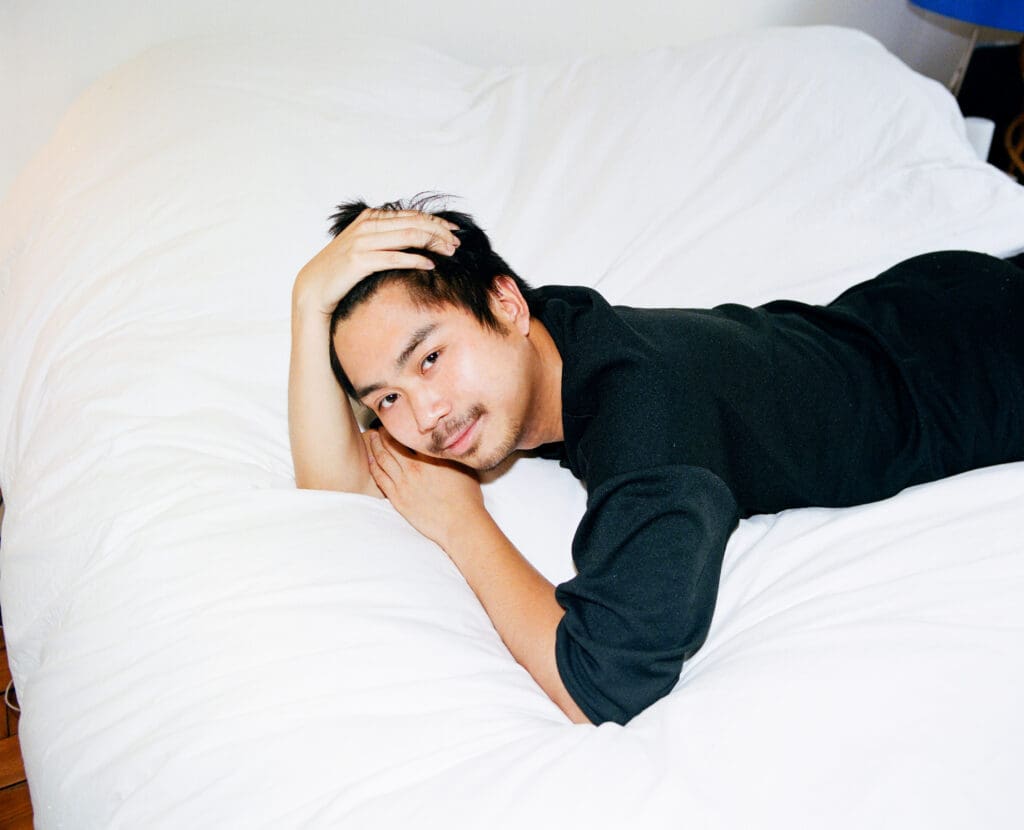 A smiling Asian man lies on his stomach on a white bed with one hand running through his hair and the other under his cheek.
