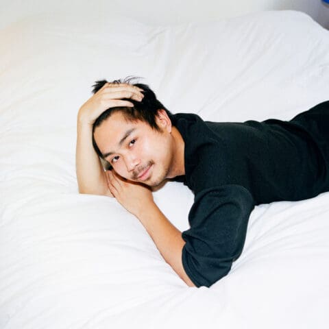 A smiling Asian man lies on his stomach on a white bed with one hand running through his hair and the other under his cheek.