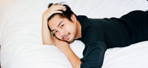 A smiling Asian man lies on his stomach on a white bed with one hand running through his hair and the other under his cheek.