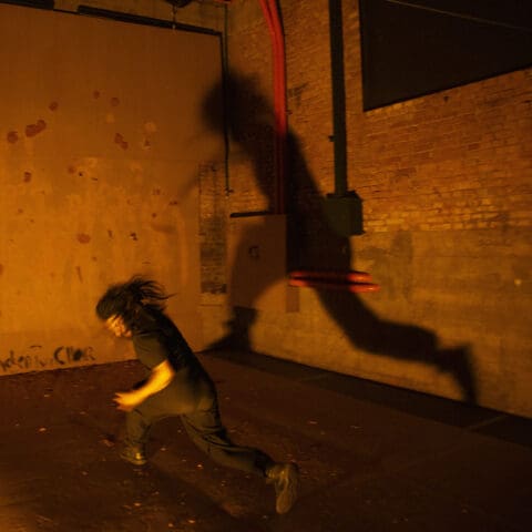 A blurry figure in all black runs across a room lit up orange with their shadow cast on the brick wall behind them.