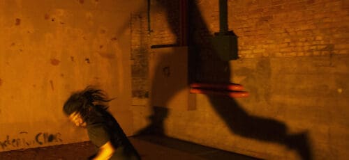 A blurry figure in all black runs across a room lit up orange with their shadow cast on the brick wall behind them.