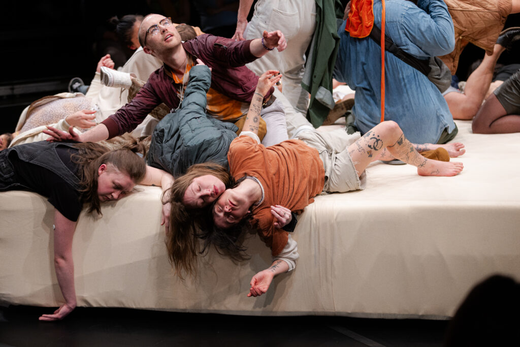 Five people lie haphazardly on a large, white platform with their limbs splayed out while one man sits halfway up reaching out.