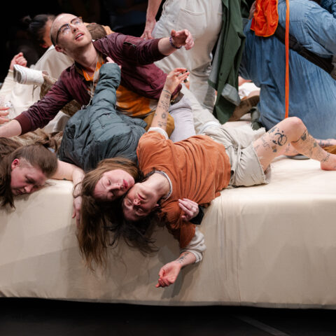 Five people lie haphazardly on a large, white platform with their limbs splayed out while one man sits halfway up reaching out.