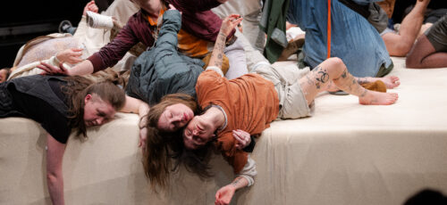 Five people lie haphazardly on a large, white platform with their limbs splayed out while one man sits halfway up reaching out.
